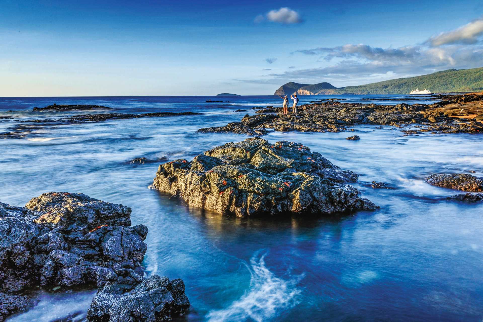 Travelers take photos on lava rocks on Santiago Island, Galápagos with the ship National Geographic Endeavour II in the background.