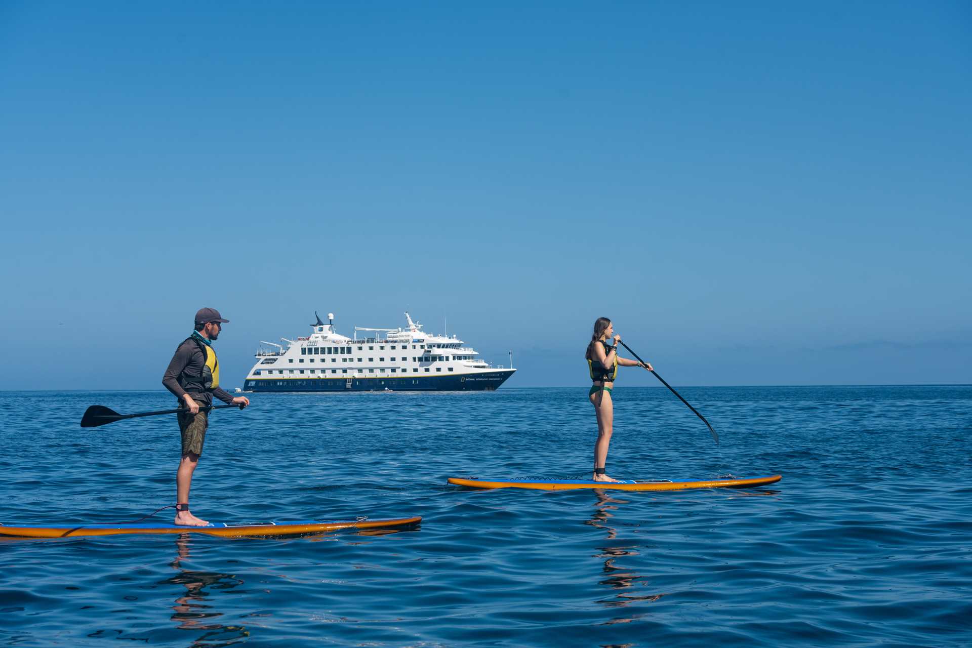 Travelers paddle on standup paddleboards off Santiago Island, Galápagos with the ship National Geographic Endeavour II in the distance.